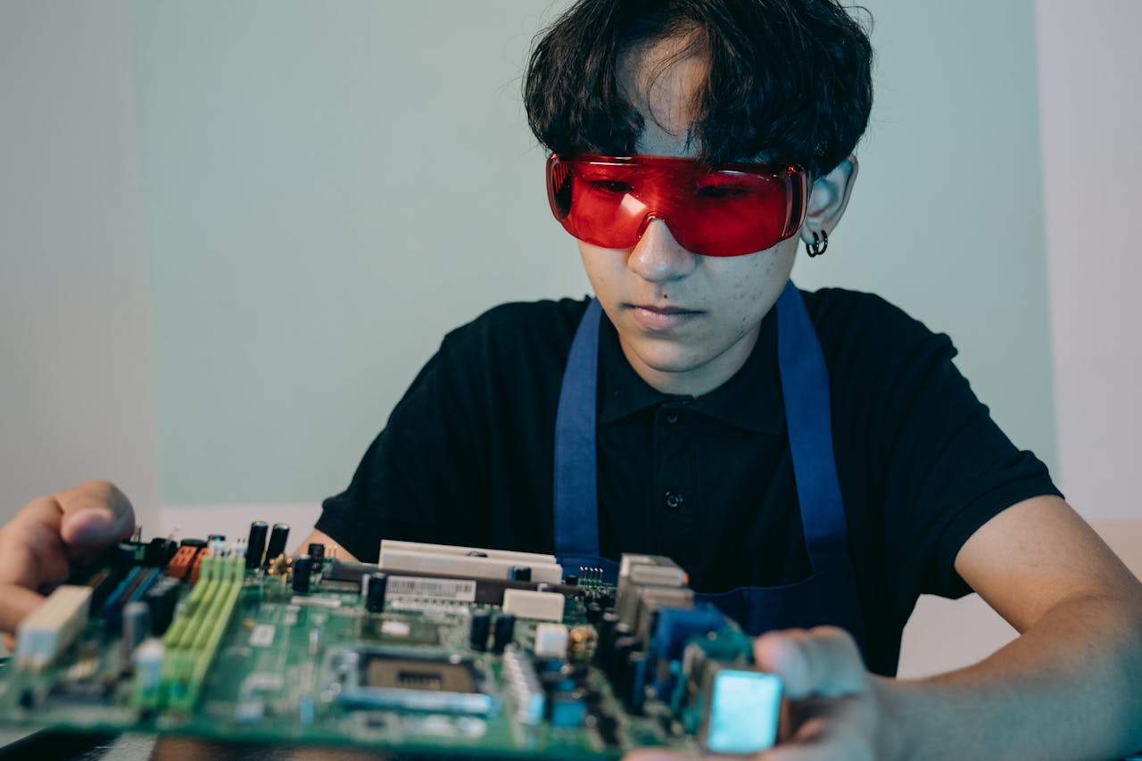 pexels photo 9242263 A young technician with red goggles examines a computer circuit board in a workshop setting.